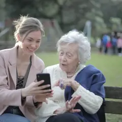 Młoda kobieta pokazuje coś na telefonie starszej pani. To scena z życia, jakby z kursu opiekuna osoby starszej, gdzie uczą się nowych technologii.