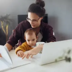 Pracująca mama z dzieckiem na kolanach analizuje dokumenty, zastanawiając się, ile zarabia opiekunka na godzinę.