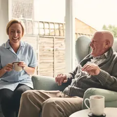 Uśmiechnięta terapeutka zajęciowa z kawą rozmawia z seniorem. Dobre zarobki terapeuty zajęciowego to satysfakcja z pracy.