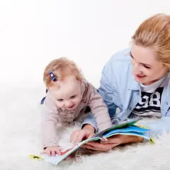 Opiekunka z kwalifikacjami do żłobka czyta książeczkę dziecku, które raczkuje na białym dywanie.