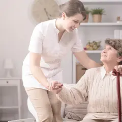 Uśmiechnięta pielęgniarka środowiskowa pomaga starszej kobiecie wstać. Wskazówki, jak załatwić opiekę.