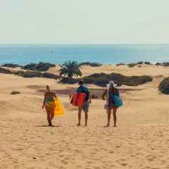 Trzy osoby idą przez wydmy na Wyspach Kanaryjskich, niosąc sprzęt plażowy. Euro to waluta, którą tu zapłacisz.