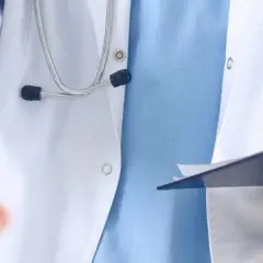 Lekarz w białym kitlu z niebieską koszulą i stetoskopem wyciąga rękę do uścisku, trzymając teczkę. To obraz pracy opiekuna medycznego w hospicjum.