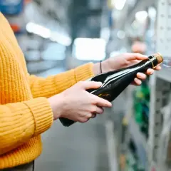Osoba w żółtym swetrze wybiera butelkę wina w sklepie. Sprawdza ceny alkoholu na Słowacji, porównując dostępne opcje.