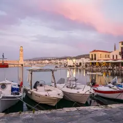 Port na Krecie o zachodzie słońca. Łodzie cumują przy nabrzeżu, a w tle widać latarnię morską i zabudowania. Kreta waluta to nie tylko pieniądze, ale i takie widoki.