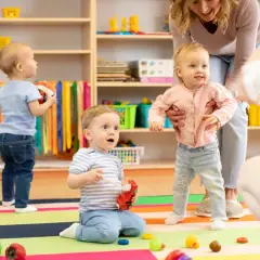 Starszy opiekun w żłobku po latach radości z maluchami. Dzieci bawią się zabawkami na kolorowej macie, a opiekunka pomaga dziewczynce wstać.