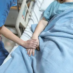 Opiekunka trzyma za rękę pacjentkę w hospicjum dla osób starszych. Ciepło i troska w tym trudnym miejscu.