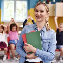 Uśmiechnięta prowadząca kurs opiekunki dziecięcej trzyma zeszyty. W tle dzieci z podniesionymi rękami.