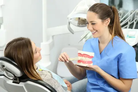 Uśmiechnięta higienistka stomatologiczna pokazuje pacjentce, jak prawidłowo szczotkować zęby, używając modelu szczęki. Dyskutują o tym, ile zarabia higienistka stomatologiczna.