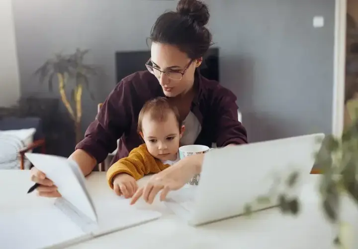 Pracująca mama z dzieckiem na kolanach analizuje dokumenty, zastanawiając się, ile zarabia opiekunka na godzinę.