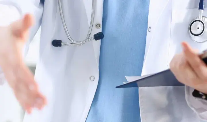 Lekarz w białym kitlu z niebieską koszulą i stetoskopem wyciąga rękę do uścisku, trzymając teczkę. To obraz pracy opiekuna medycznego w hospicjum.