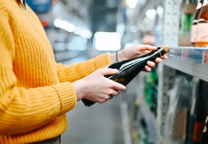 Osoba w żółtym swetrze wybiera butelkę wina w sklepie. Sprawdza ceny alkoholu na Słowacji, porównując dostępne opcje.