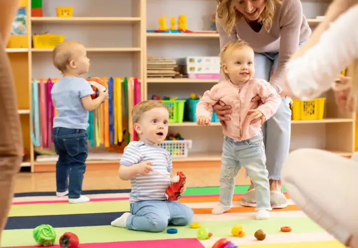 Starszy opiekun w żłobku po latach radości z maluchami. Dzieci bawią się zabawkami na kolorowej macie, a opiekunka pomaga dziewczynce wstać.