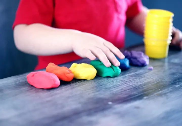 Dziecko w czerwonej koszulce bawi się plasteliną w tęczowych kolorach. To idealny obrazek do materiałów o kursie na opiekuna w żłobku.