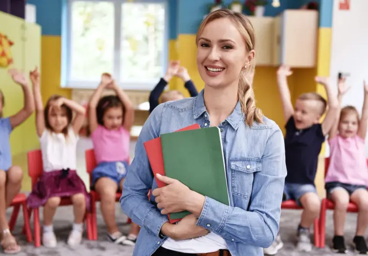Uśmiechnięta prowadząca kurs opiekunki dziecięcej trzyma zeszyty. W tle dzieci z podniesionymi rękami.