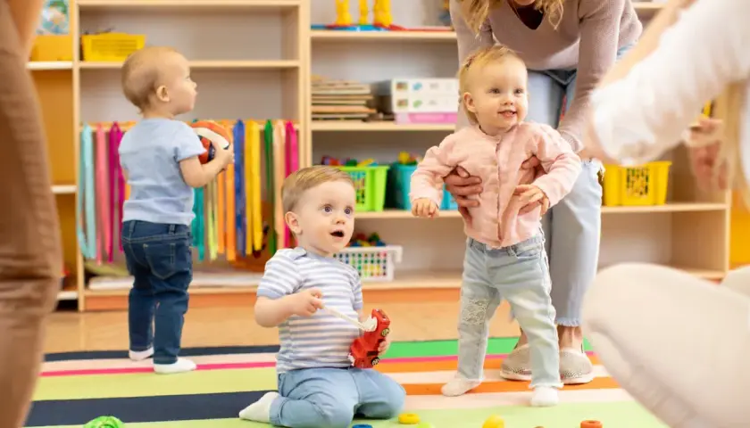 Starszy opiekun w żłobku po latach radości z maluchami. Dzieci bawią się zabawkami na kolorowej macie, a opiekunka pomaga dziewczynce wstać.