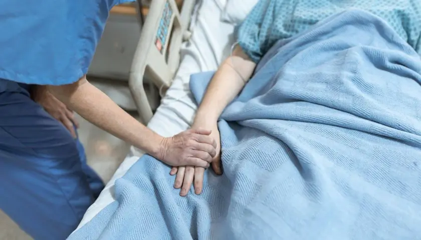 Opiekunka trzyma za rękę pacjentkę w hospicjum dla osób starszych. Ciepło i troska w tym trudnym miejscu.