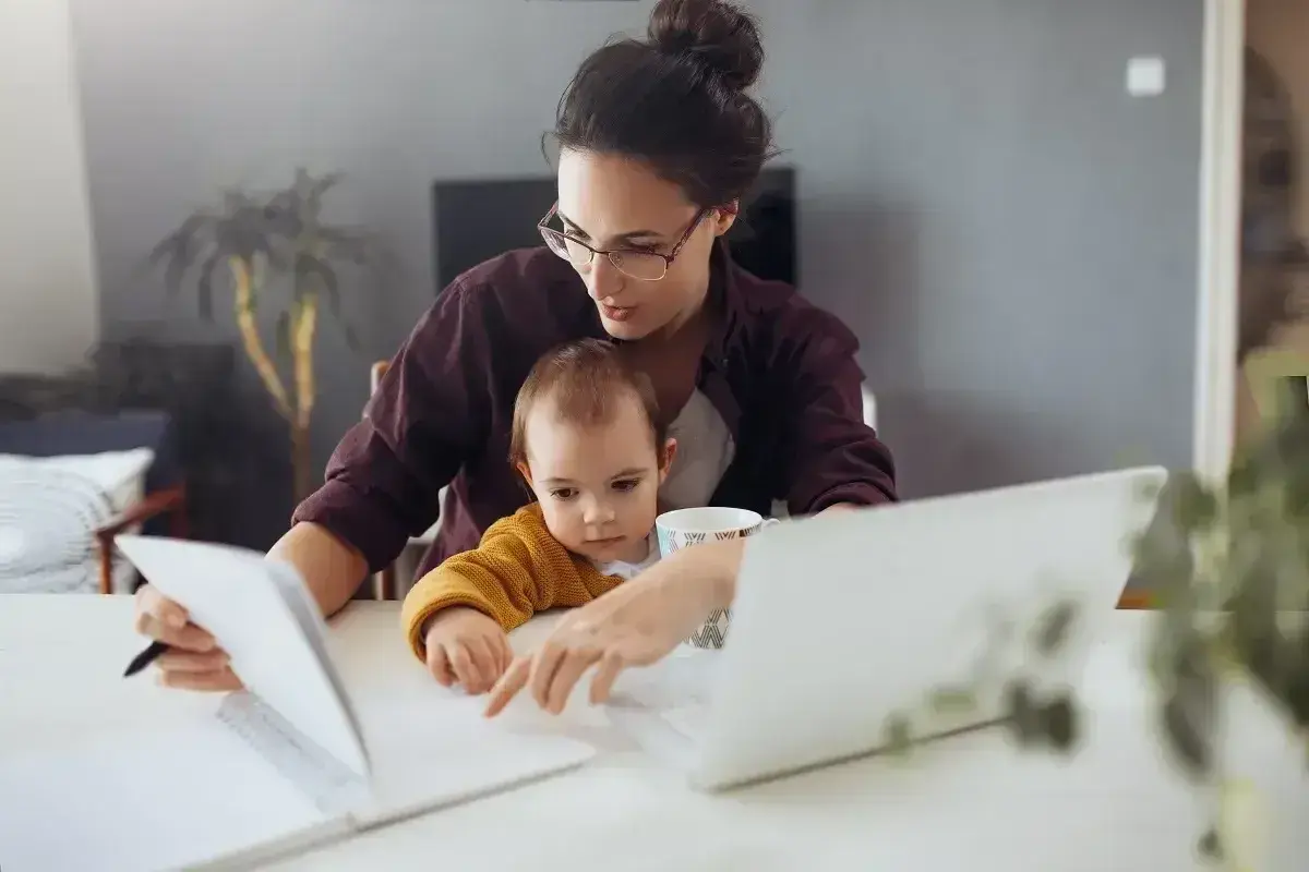 Pracująca mama z dzieckiem na kolanach analizuje dokumenty, zastanawiając się, ile zarabia opiekunka na godzinę.