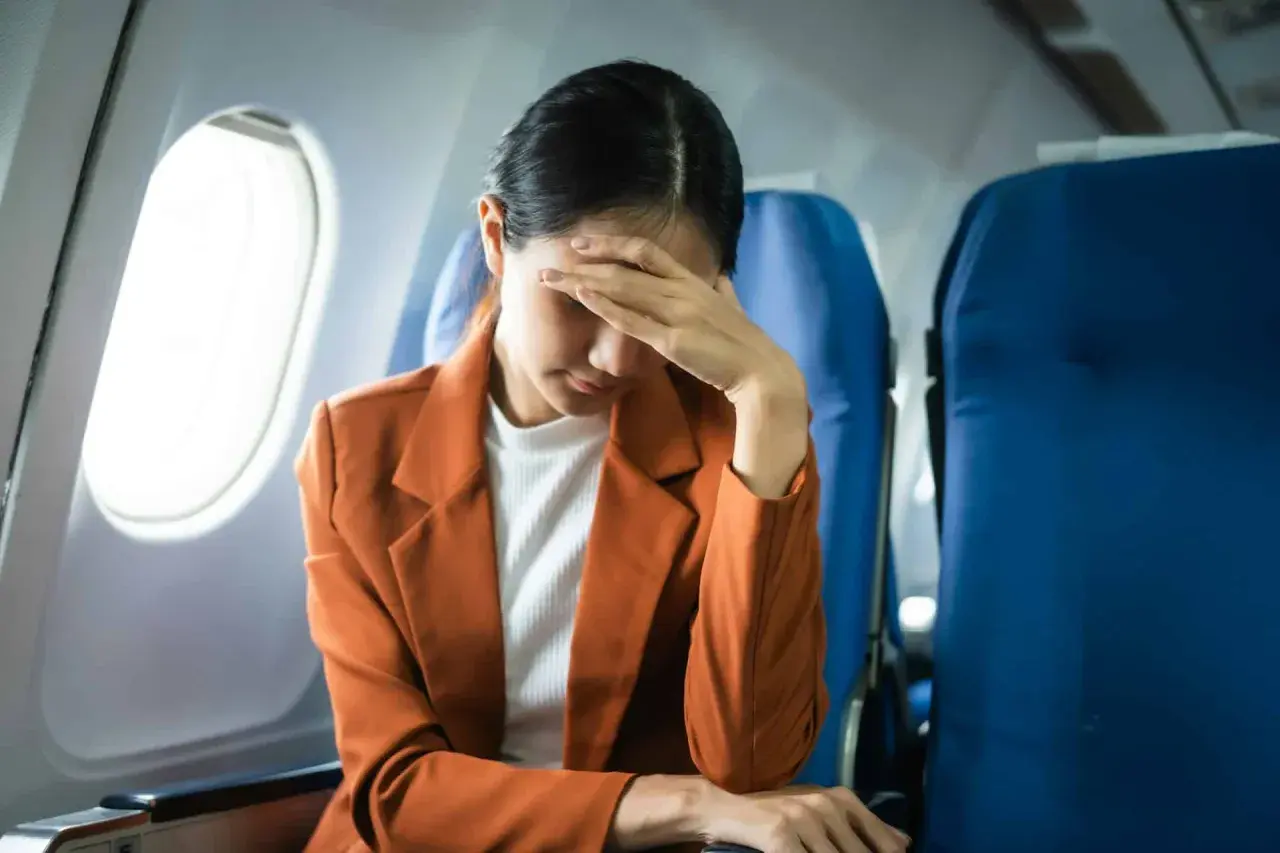 Kobieta w samolocie z ręką na czole, wyraźnie cierpiąca na jet lag. Co to jest jet lag? To zmęczenie po podróży.