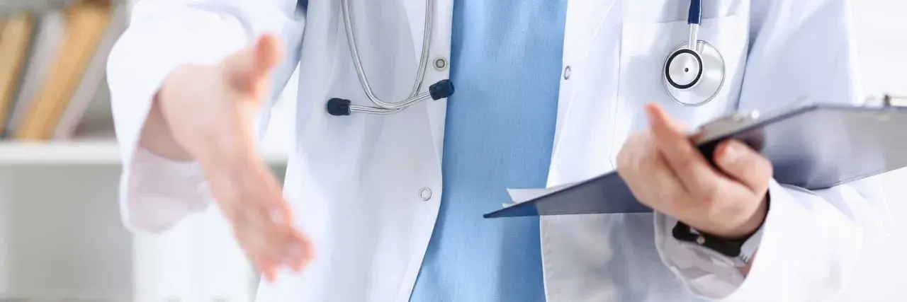 Lekarz w białym kitlu z niebieską koszulą i stetoskopem wyciąga rękę do uścisku, trzymając teczkę. To obraz pracy opiekuna medycznego w hospicjum.