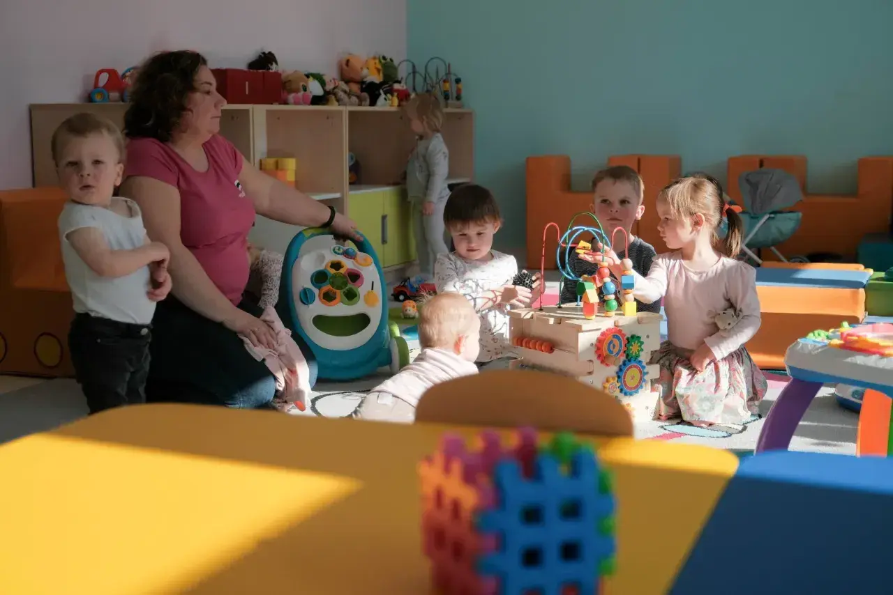 Jedna opiekunka w żłobku dogląda kilkorga dzieci bawiących się zabawkami.