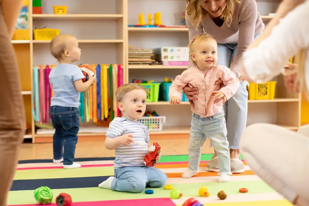 Starszy opiekun w żłobku po latach radości z maluchami. Dzieci bawią się zabawkami na kolorowej macie, a opiekunka pomaga dziewczynce wstać.