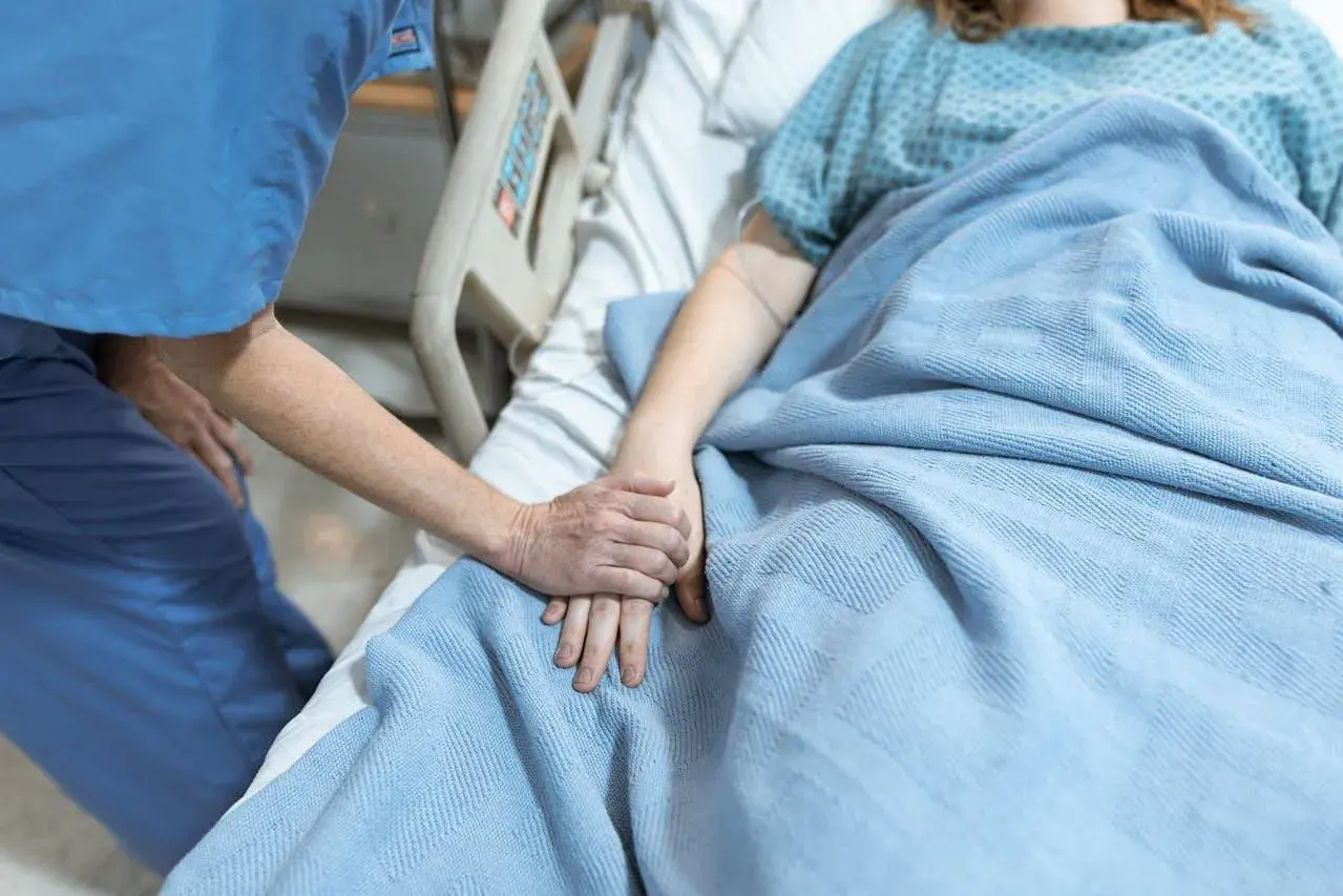 Opiekunka trzyma za rękę pacjentkę w hospicjum dla osób starszych. Ciepło i troska w tym trudnym miejscu.