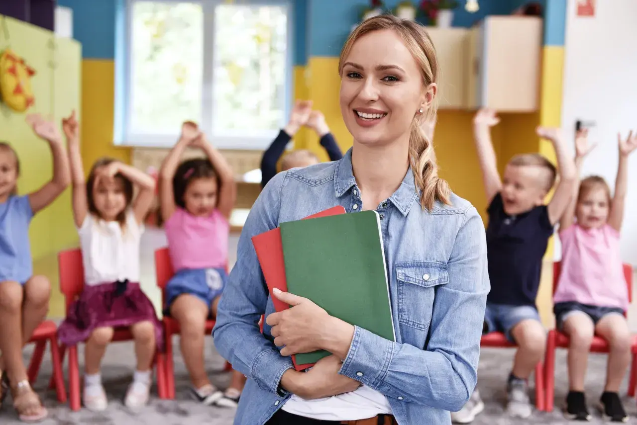 Uśmiechnięta prowadząca kurs opiekunki dziecięcej trzyma zeszyty. W tle dzieci z podniesionymi rękami.