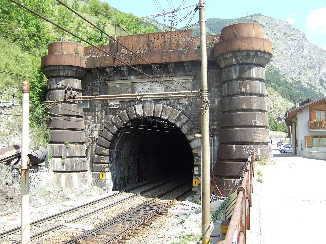 Kamienne filary i łuk wejściowy do tunelu Frejus, z widocznymi torami kolejowymi i g&oacute;rami w tle.