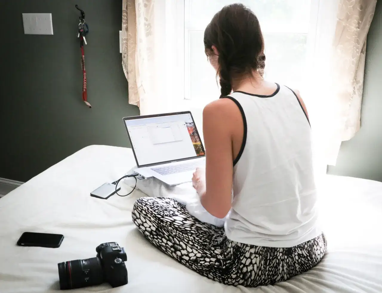 Kobieta pracuje na laptopie, siedząc na łóżku. Wygląda jak terapeuta zajęciowy online, przygotowując materiały do sesji.