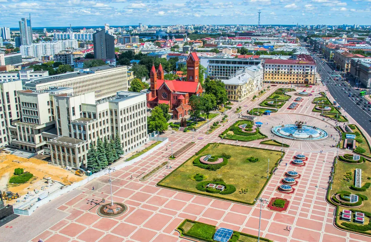 Plac w Mińsku z czerwonym kościołem i fontanną. W tle nowoczesne budynki, widok na miasto. Ceny na Białorusi mogą być r&oacute;żne w zależności od lokalizacji.
